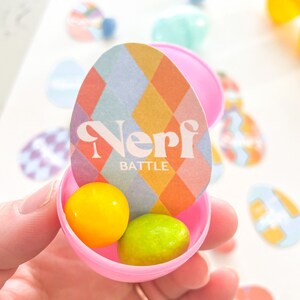 Könnte beinhalten: Rosa Plastik-Osterei mit einem bunten gemusterten Papierüberzug, auf dem "Nerf Battle" steht. Das Ei enthält zwei kleine Süßigkeiten, eine gelbe und eine grüne.