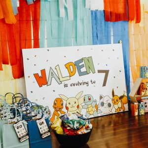 May include: A white sign with the name "Walden" in colourful letters and the number "7" next to it. The text "is evolving to" is written below the name. The sign features cartoon images of Pokemon characters. There are blue and green gift bags with Pokemon character tags on a wooden table.