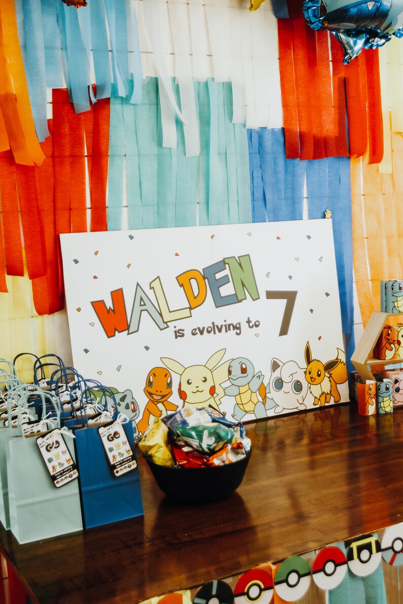 May include: A white sign with the name "Walden" in colourful letters and the number "7" next to it. The text "is evolving to" is written below the name. The sign features cartoon images of Pokemon characters. There are blue and green gift bags with Pokemon character tags on a wooden table.