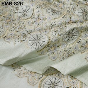 May include: Pale green fabric with intricate embroidery. The design features repeating circular patterns with radiating lines and floral accents, in shades of silver, gold, and pale purple. The fabric is folded, showing texture and detail. The text "EMB-826" is visible.
