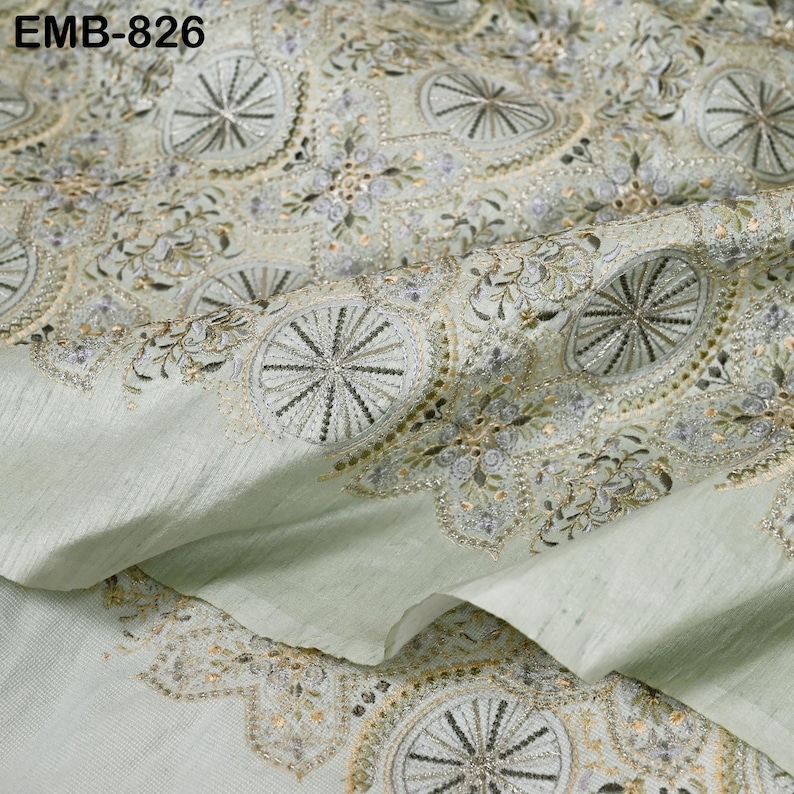 May include: Pale green fabric with intricate embroidery. The design features repeating circular patterns with radiating lines and floral accents, in shades of silver, gold, and pale purple. The fabric is folded, showing texture and detail. The text "EMB-826" is visible.