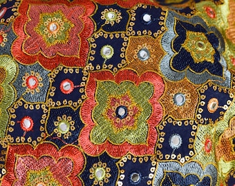 May include: A close-up of a dark blue fabric with intricate embroidery in shades of red, green, gold, and silver. The fabric is adorned with small round mirrors, creating a shimmering effect.