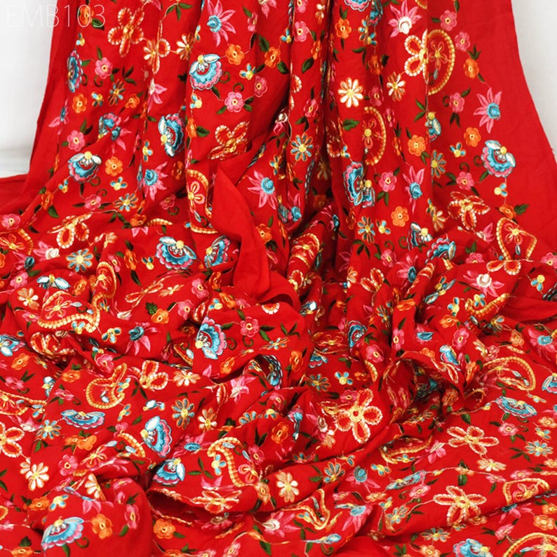 May include: Red fabric with a floral embroidery pattern. The fabric is folded and shows the intricate details of the embroidery. The flowers are in shades of pink, blue, yellow, and green.