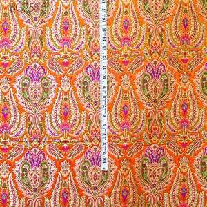 Op de afbeelding: Oranje stof met een gouden en paarse paisley-print. De stof wordt gemeten met een liniaal in inches.