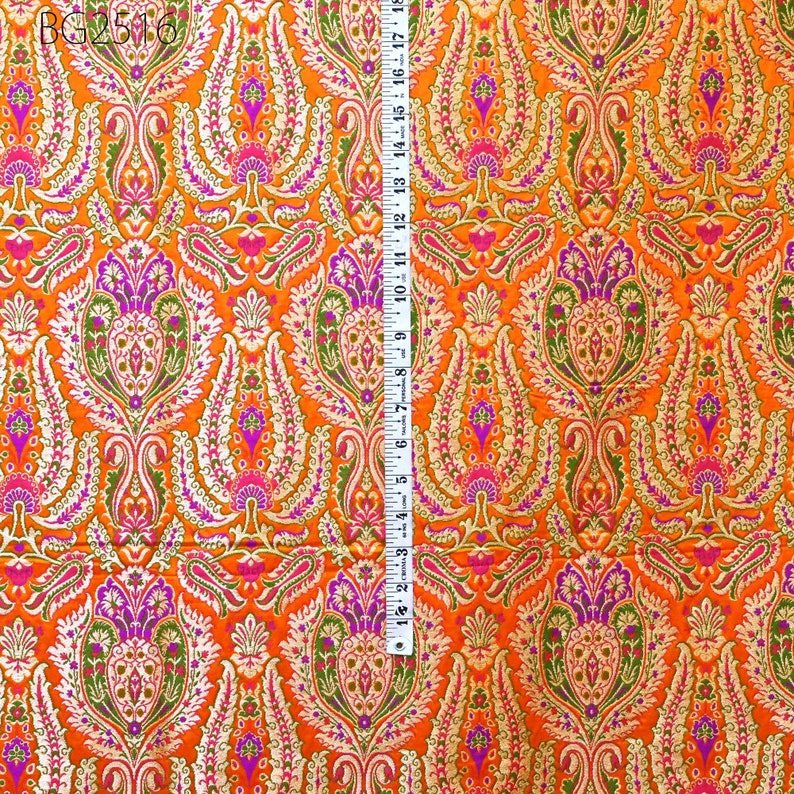 Op de afbeelding: Oranje stof met een gouden en paarse paisley-print. De stof wordt gemeten met een liniaal in inches.