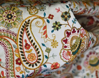 Op de afbeelding: Witte stof met kleurrijke paisley- en bloemenborduurwerk. De stof heeft een herhalend patroon van paisley-vormen, bloemen en bladeren in tinten rood, oranje, geel, groen, blauw en zwart.