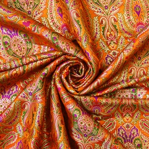 Op de afbeelding: Oranje stof met een paisley-patroon in goud, groen en paars. De stof is gevouwen en gedrapeerd om het patroon te laten zien.