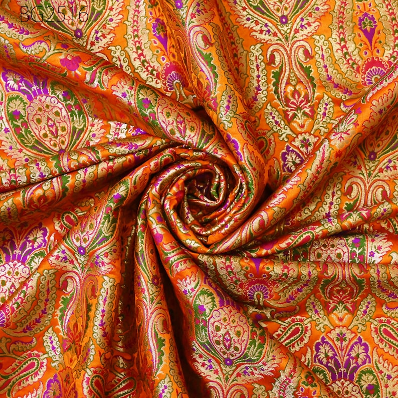 Op de afbeelding: Oranje stof met een paisley-patroon in goud, groen en paars. De stof is gevouwen en gedrapeerd om het patroon te laten zien.