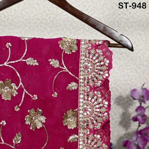 Puede incluir: Una vibrante tela fucsia con intrincados bordados florales dorados y detalles de lentejuelas. El borde presenta un patr&oacute;n decorativo con detalles de espejo. El texto "ST-948" es visible. La tela est&aacute; colgada de una percha de madera.