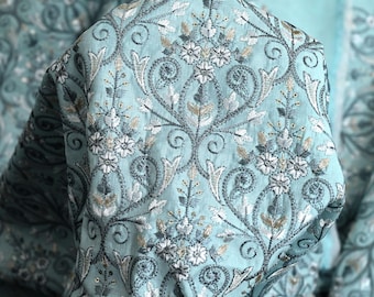 May include: A close-up of a light blue fabric with a white and silver floral embroidery pattern. The fabric has a subtle texture and a delicate, intricate design.