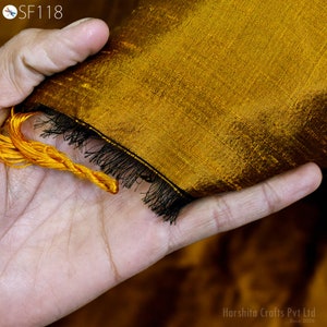 May include: A close-up of a gold fabric with a black fringe trim. The fabric is being held in a hand.