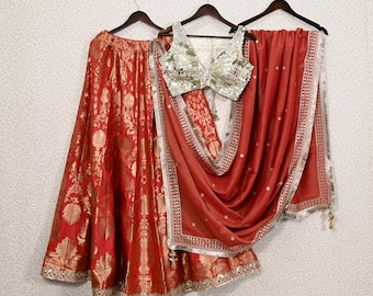 May include: A three-piece Indian ethnic wear set. The set includes a red skirt with gold floral patterns, a silver and gold embellished top, and a red shawl with silver trim. The outfit is displayed on hangers against a textured wall.