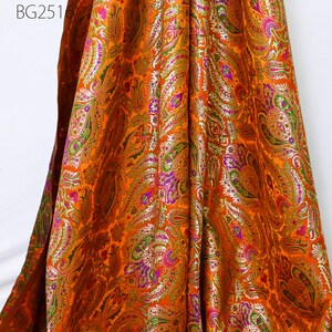 Op de afbeelding: Oranje stof met een paisley-patroon in goud, paars, groen en roze. De stof heeft een metallic glans.