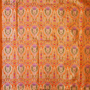 Op de afbeelding: Oranje stof met een herhalend patroon van gouden, groene, roze en paarse bloemenmotieven. De stof heeft een subtiele glans.