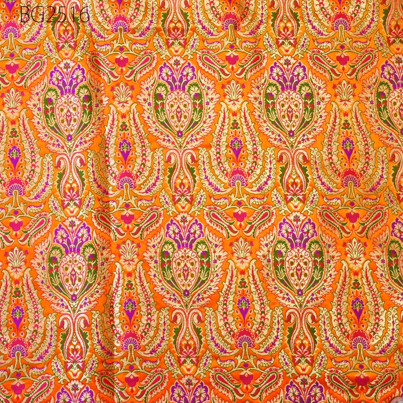 Op de afbeelding: Oranje stof met een herhalend patroon van gouden, roze en groene bloemenmotieven. De stof heeft een brokaattextuur.
