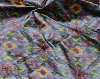 May include: A close-up of a blue fabric with a colorful ikat pattern. The fabric has a subtle sheen and is woven with a variety of colors, including red, green, yellow, and black.