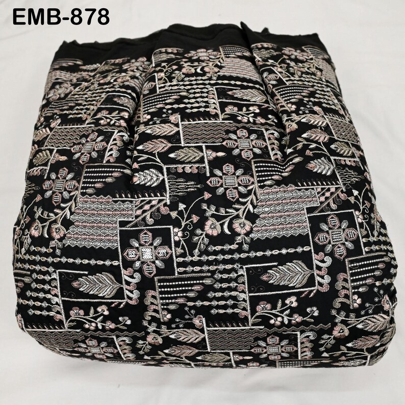 May include: Black fabric featuring an embroidered floral and geometric pattern with pink, white, and silver accents. The text "EMB-878" is visible. The fabric is folded, suggesting it is a textile, possibly for clothing or home decor.