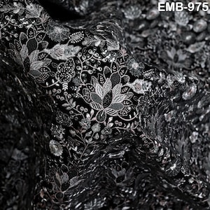 May include: Close-up of black fabric with detailed floral embroidery in silver and pale pink thread. The design features leaves, flowers, and vines. The text "EMB-975" is visible. This textile is suitable for clothing and home decor projects.