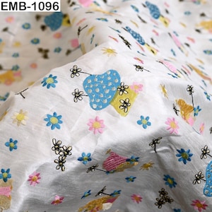 May include: White fabric featuring an embroidered pattern of blue, yellow, and pink shapes alongside small multi-colored flowers and black line-art floral motifs. The text EMB-1096 is visible in the top left corner of the fabric material.