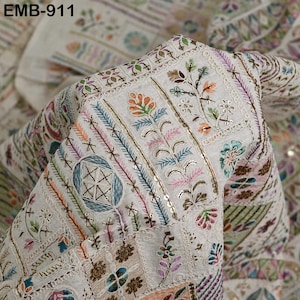 May include: Close-up of a cream fabric with detailed embroidery. The design incorporates floral and geometric patterns in colours such as orange, blue, pink, and green. The fabric has a textured look with the text "EMB-911" visible.