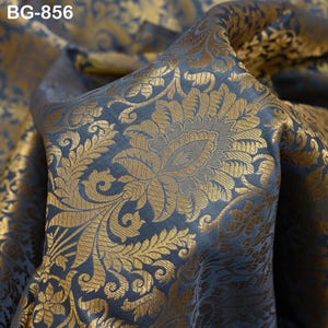 Pode incluir: Close-up de um tecido luxuoso com um padrão floral. O tecido é azul escuro com detalhes dourados, criando um visual rico e elegante. O design apresenta uma grande flor ornamentada no centro, rodeada por folhas e vinhas em redemoinho. O texto "BG-856" está no canto superior esquerdo.