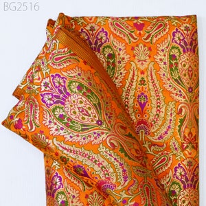 Op de afbeelding: Oranje stof met een paisley-patroon in tinten paars, groen en goud. De stof is gevouwen en heeft een gouden rand.