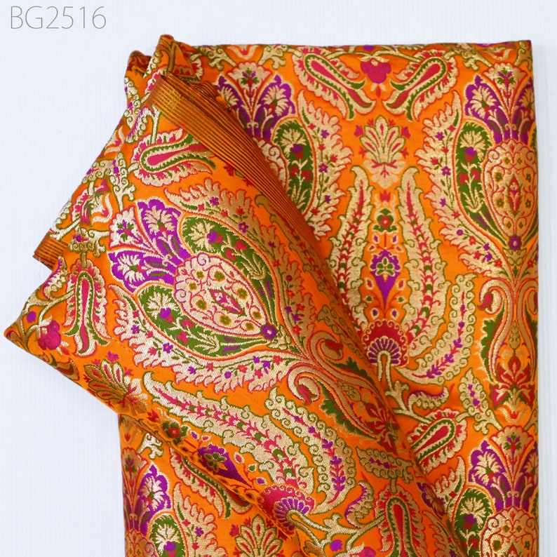 Op de afbeelding: Oranje stof met een paisley-patroon in tinten paars, groen en goud. De stof is gevouwen en heeft een gouden rand.