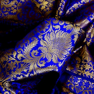 以下が含まれることがあります： 金色の織り模様が施された青い布。布は折り畳まれ、ドレープされており、模様の複雑なディテールがわかります。