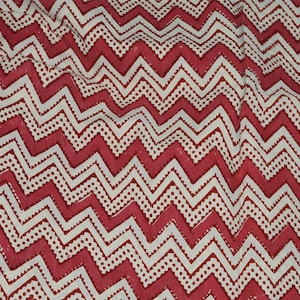 May include: Close-up of a fabric with a repeating zigzag pattern in red and white. The design features alternating rows of solid zigzags and dotted lines, creating a textured appearance. The fabric appears to be cotton or a similar material.