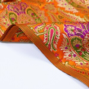 Op de afbeelding: Oranje stof met een paisley-patroon in tinten groen, paars, goud en roze. De stof heeft een gouden en groene rand.