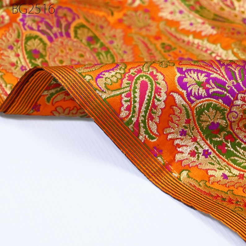 Op de afbeelding: Oranje stof met een paisley-patroon in tinten groen, paars, goud en roze. De stof heeft een gouden en groene rand.
