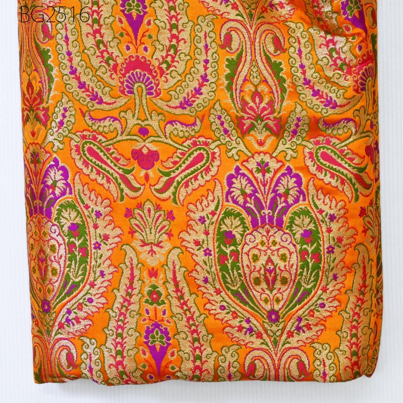 Op de afbeelding: Oranje stof met een paisley-patroon in goud, groen, roze en paars.