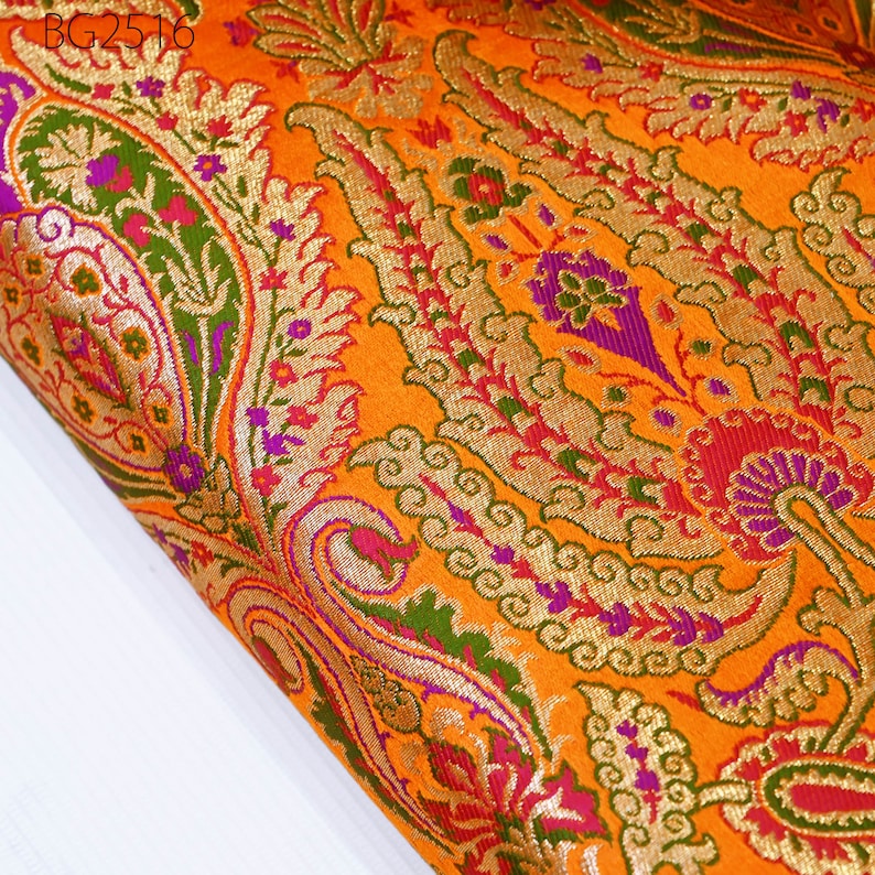 Op de afbeelding: Oranje stof met een paisley-patroon in goud, groen, paars en rood. De stof heeft een geweven textuur en een glanzende afwerking.