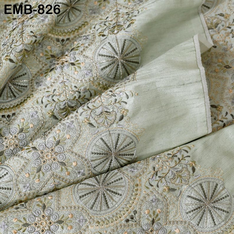 May include: Pale green fabric with intricate embroidery. The design features repeating circular patterns with radial spokes and floral accents in shades of gold, silver, and green. The text "EMB-826" is visible in the upper left corner. The fabric has a subtle sheen.