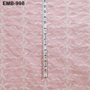 Op de afbeelding: Roze stof met witte bloemenborduurwerk. Een meetlint geeft aan dat de stof ongeveer 35,5 cm breed is. De tekst "EMB-998" staat in de linkerbovenhoek. De stof heeft een delicate, getextureerde uitstraling.