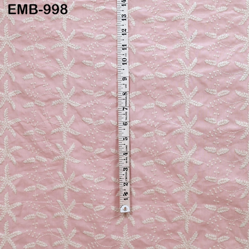 Op de afbeelding: Roze stof met witte bloemenborduurwerk. Een meetlint geeft aan dat de stof ongeveer 35,5 cm breed is. De tekst "EMB-998" staat in de linkerbovenhoek. De stof heeft een delicate, getextureerde uitstraling.