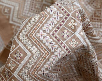 May include: Close-up of a tan fabric with intricate embroidery. The design features geometric patterns, floral motifs, and the text "EMB-859" in the upper left corner. The embroidery incorporates white, peach, and burgundy threads.