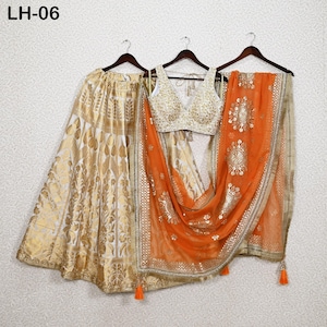 Peut inclure: Un ensemble de vêtements ethniques indiens en trois pièces. L'ensemble comprend une jupe à motifs or et blanc, un haut brodé blanc et or et une écharpe orange transparente avec des embellissements dorés. Le texte "LH-06" est visible.