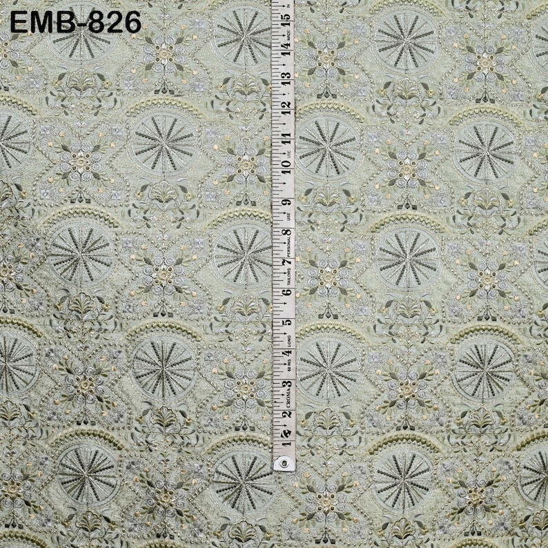 May include: A light green fabric with an intricate embroidered pattern featuring circular and floral designs. A ruler is placed vertically along the fabric, showing measurements in inches. The text "EMB-826" is visible in the upper left corner.