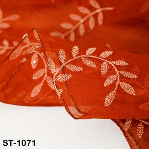 以下が含まれることがあります： 繊細な白い葉の模様が刺繍された、透明感のある錆色の生地のクローズアップ。生地は折りたたまれ、その質感と刺繍の複雑なディテールが明らかになっています。「ST-1071」の文字が下部に表示されています。