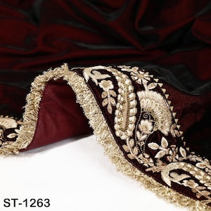 May include: Close-up of a burgundy fabric with intricate gold embroidery and a gold fringe trim. The fabric has a velvet-like texture and features floral and leaf patterns. The text "ST-1263" is visible at the bottom.