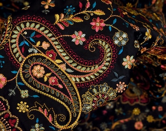 Op de afbeelding: Zwarte stof met ingewikkelde paisley-borduurwerk in goud, roze en blauw. De stof is gevouwen en toont een close-up van de gedetailleerde steken.
