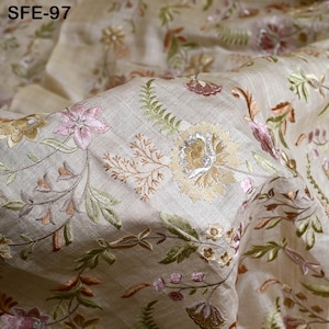 May include: Beige fabric with detailed floral embroidery in pink, green, and gold. The embroidery depicts flowers, leaves, and vines, creating a textured and elegant design. The fabric is slightly creased, adding depth. The text "SFE-97" is visible in the upper left corner of the image.