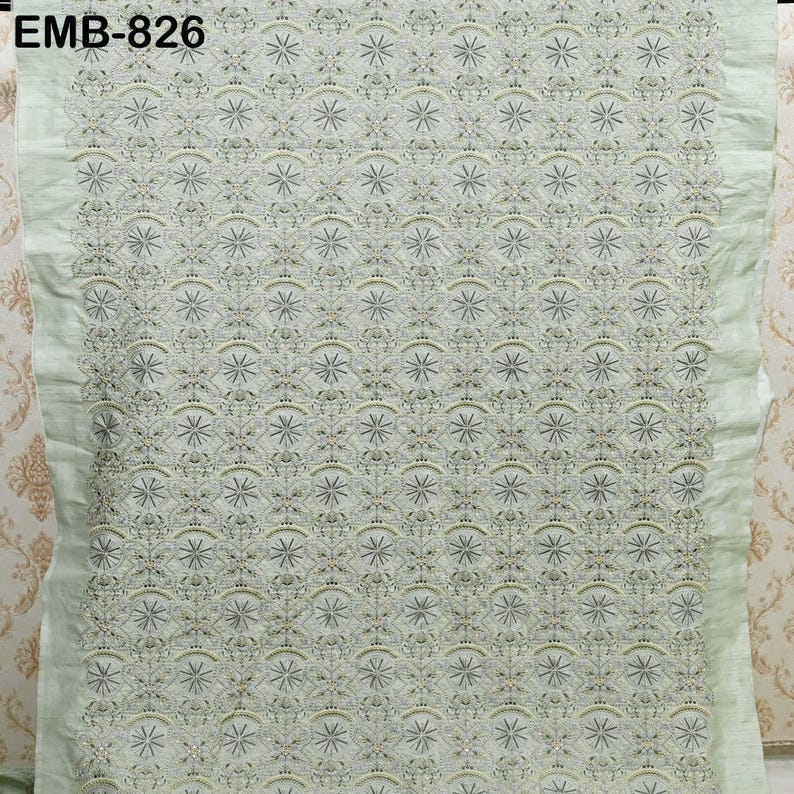 May include: A light green fabric with intricate embroidery. The design features a repeating pattern of star-like shapes and floral motifs. The text "EMB-826" is visible in the upper left corner. The fabric has a delicate, textured appearance, suitable for apparel or home decor.