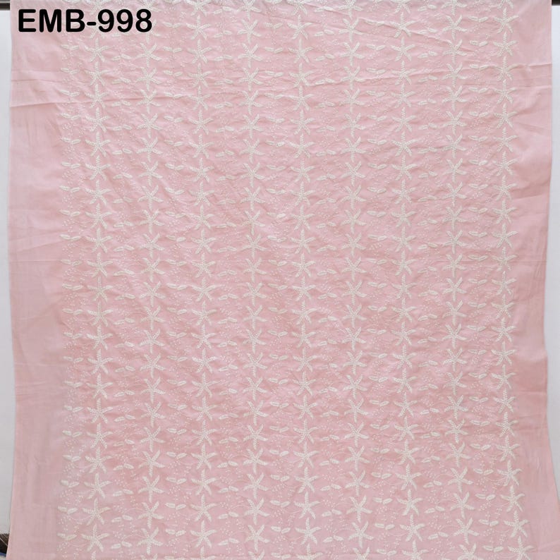 Op de afbeelding: Een lichtroze stof met een herhalend wit bloemenborduurpatroon. De tekst "EMB-998" is zichtbaar linksboven. De stof lijkt een lichtgewicht materiaal te zijn.