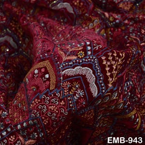 May include: Close-up of a burgundy velvet fabric with detailed embroidery. The design features floral and geometric patterns in shades of blue, gold, and white. The fabric has a luxurious sheen and is detailed with small sequins. The text "EMB-943" is visible.