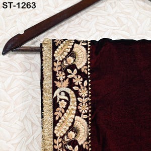 May include: A section of a maroon textile with intricate gold embroidery and a gold fringe trim. The design includes floral and paisley patterns. The textile is hanging on a wooden hanger. The text "ST-1263" is visible.