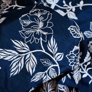 Pode incluir: Close-up de um tecido azul escuro com um padrão de bordado floral branco. O tecido tem um brilho sutil e uma textura macia.