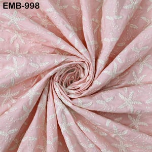 Op de afbeelding: Een close-up van een roze stof met witte bloemenborduurwerk. De stof is in een spiraal verzameld, waarbij de gedetailleerde steken en textuur worden getoond. De tekst "EMB-998" staat in de linkerbovenhoek.