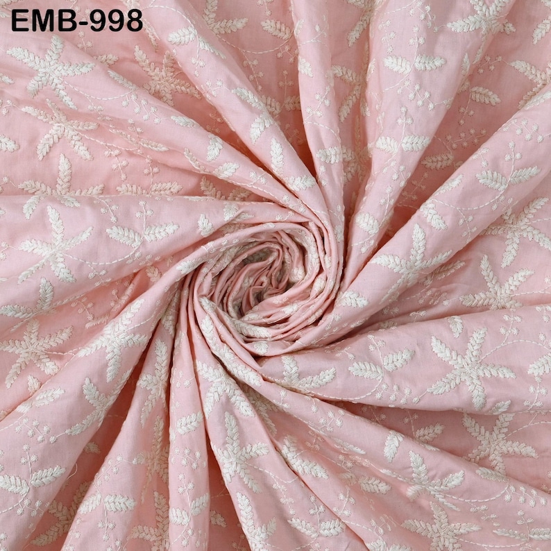Op de afbeelding: Een close-up van een roze stof met witte bloemenborduurwerk. De stof is in een spiraal verzameld, waarbij de gedetailleerde steken en textuur worden getoond. De tekst "EMB-998" staat in de linkerbovenhoek.